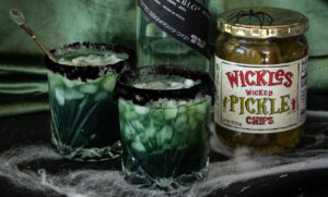 Wickles Pickles Mezcal Halloween Margaritas and a pickle jar with a spider on it. The jar and glasses are on a table with spider webs on it.